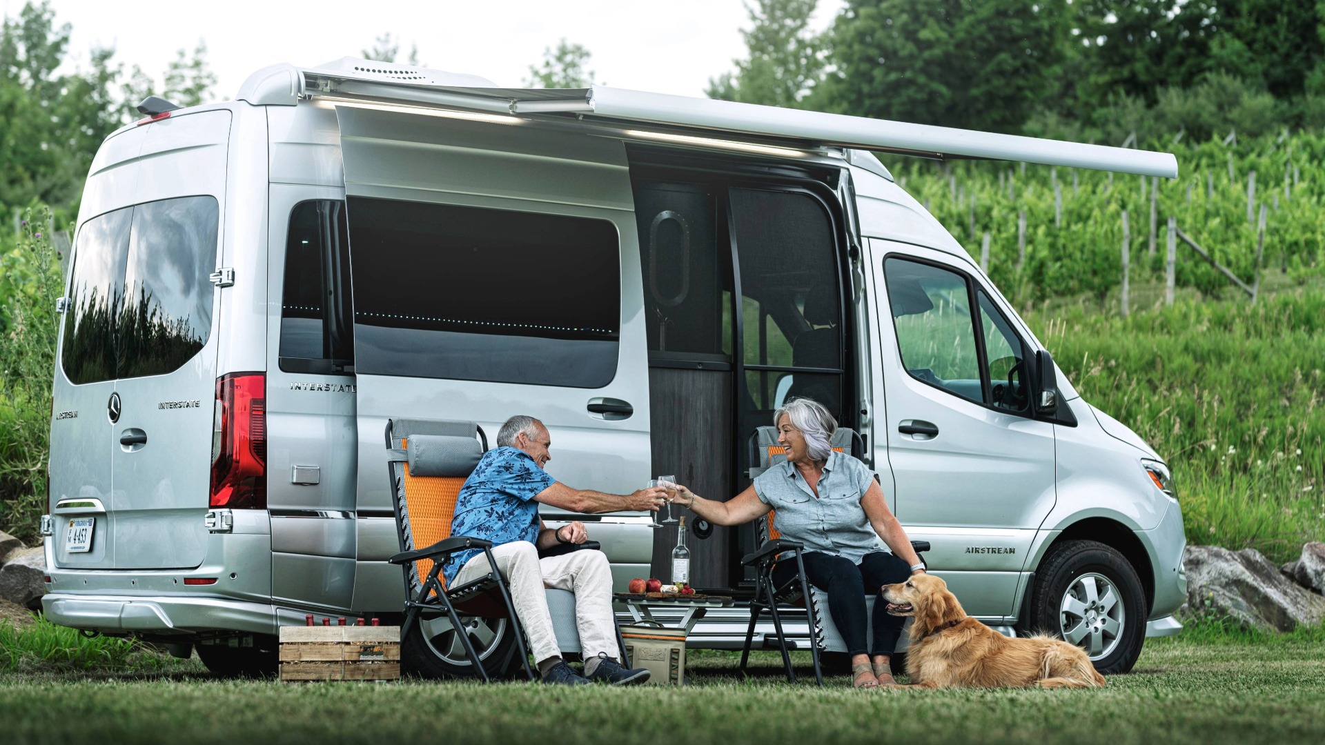 Elderly couple toasting with glasses beside a parked camper van in a grassy area, with a dog resting at their feet.