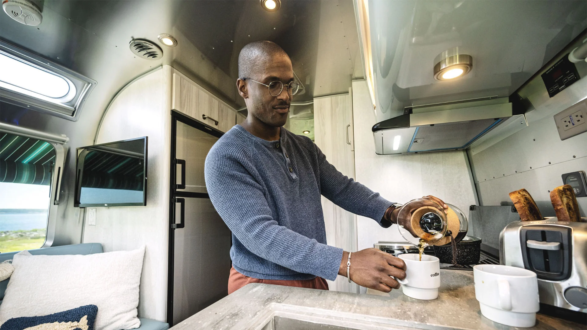 A man wearing glasses pours coffee into a mug inside a modern RV kitchen, with toast in a toaster and a TV and refrigerator in the background.