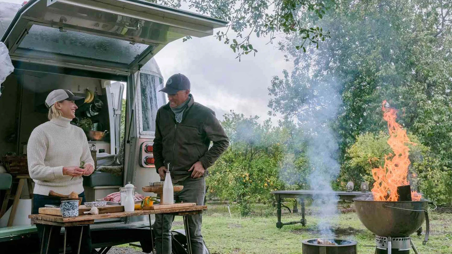 Two people stand by a camper van, preparing food on a table outdoors. A grill with flames nearby, surrounded by lush greenery.