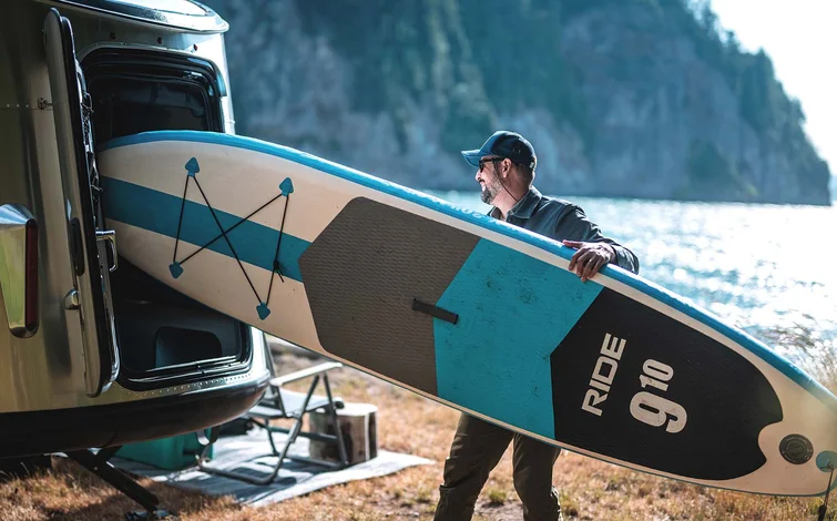 Man carrying a paddleboard towards a trailer by a lake, with trees and cliffs in the background.
