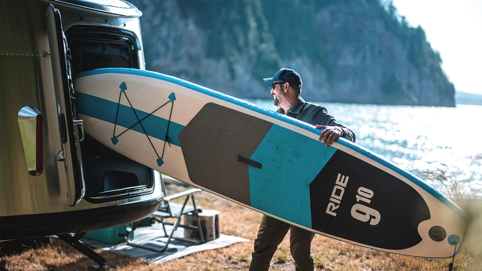 Man carrying a paddleboard towards a trailer by a lake, with trees and cliffs in the background.