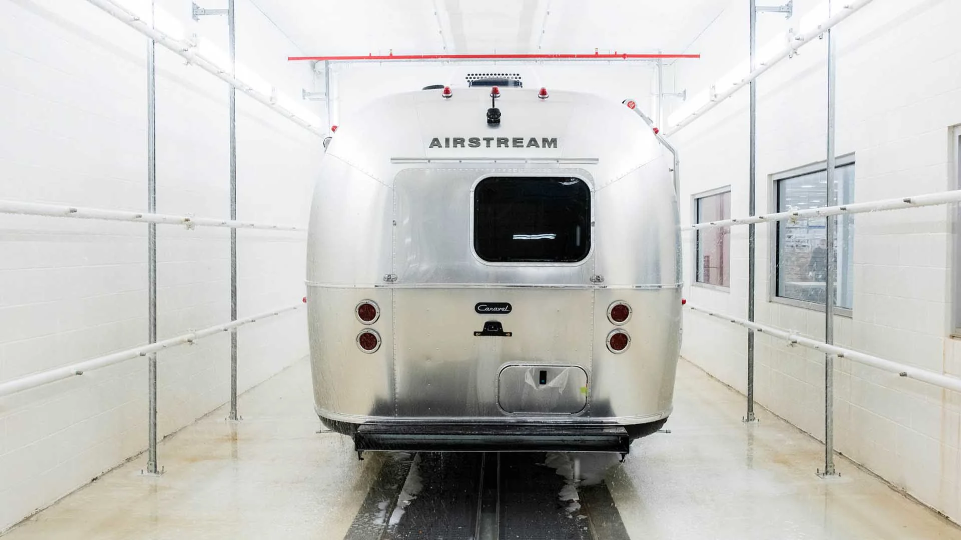 A silver Airstream trailer viewed from the rear, parked in a narrow, well-lit indoor space with white walls and piping.