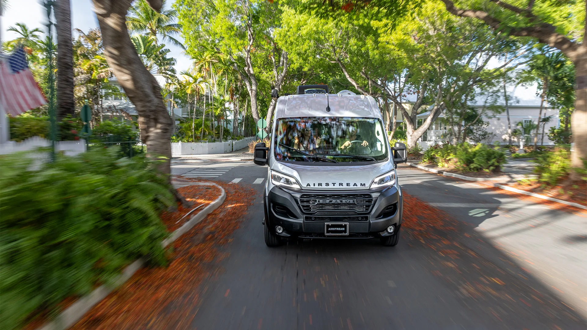 A silver Airstream camper van drives down a leafy neighborhood street, surrounded by green foliage and palm trees.