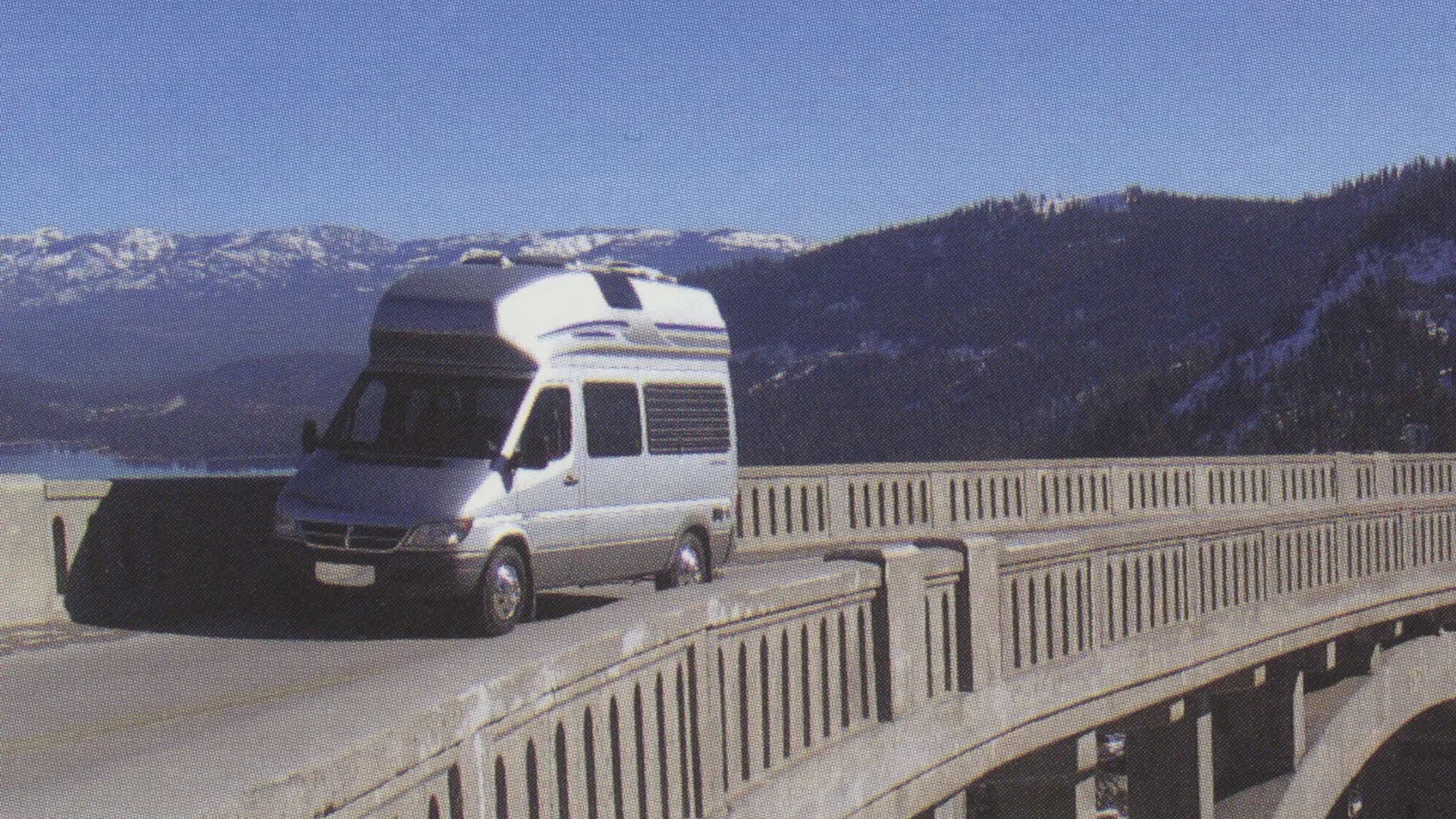 A camper van drives on a curved mountain bridge under a clear blue sky, with snow-capped peaks and forested slopes in the background.
