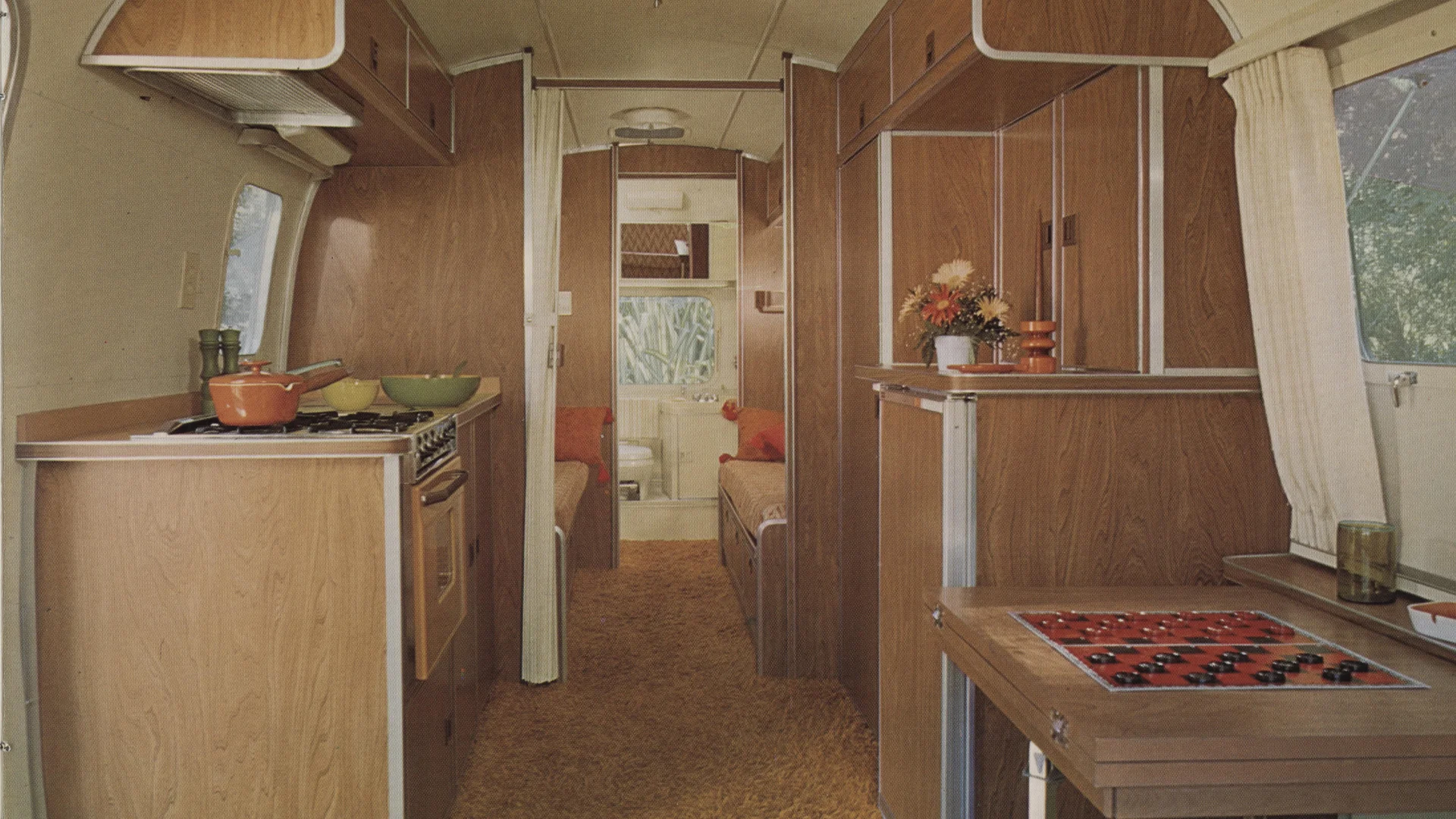 Interior of a vintage camper with wood paneling, a small kitchen on the left, two beds in the back, flowers on a shelf, and a checkers board set up on a pull-out table by the window.