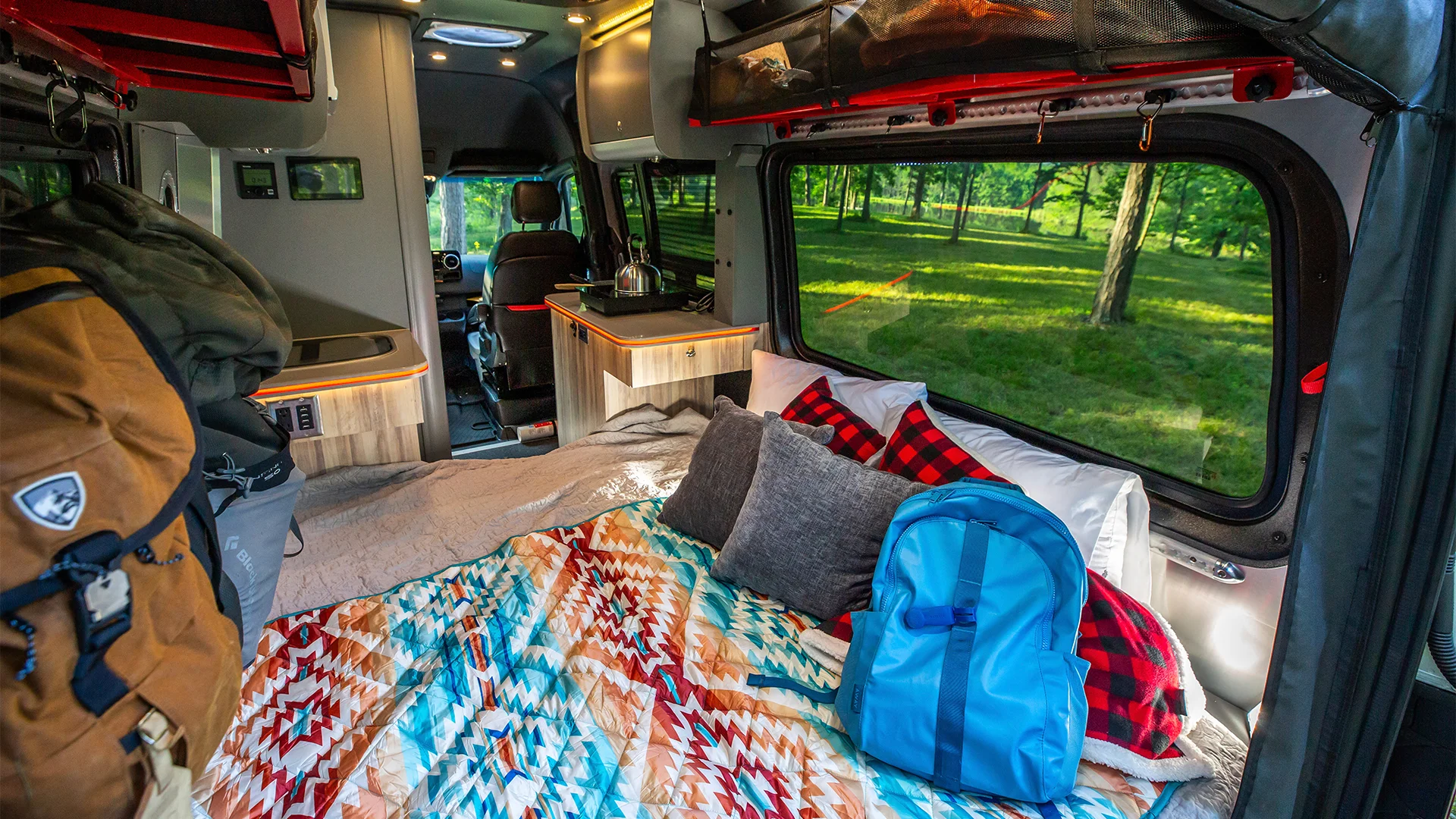 Interior of a camper van with a colorful quilted bed, pillows, backpacks, and a view of a grassy park through the large side window.