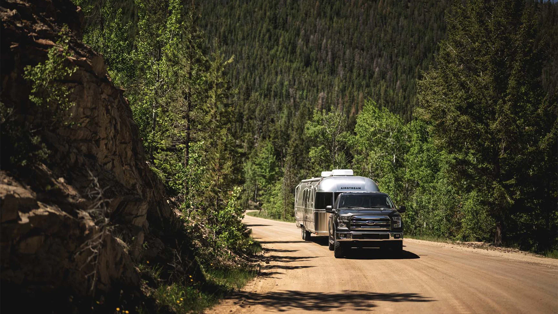 A truck towing an Airstream trailer on a forested mountain road under a clear sky, surrounded by lush green trees.