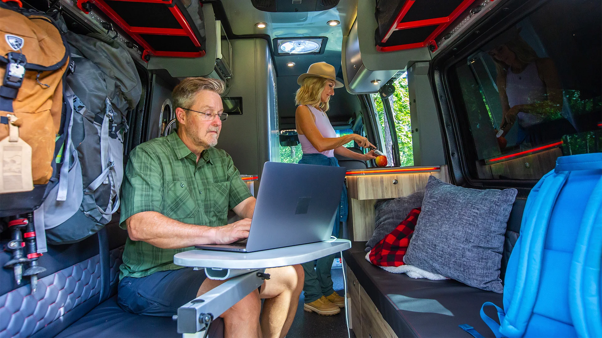 Man working on a laptop in a camper van with a fold-out table, while a woman prepares a meal in the background. Camping gear is visible.