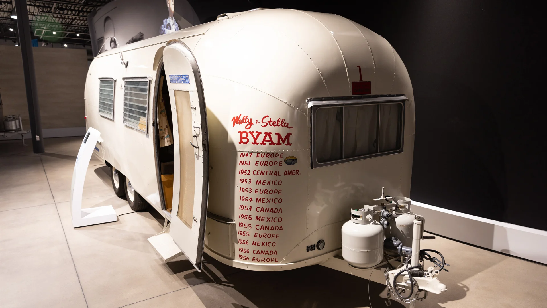 A vintage Airstream trailer on display, with travel history dates and destinations written on its side, and an open door revealing the interior.