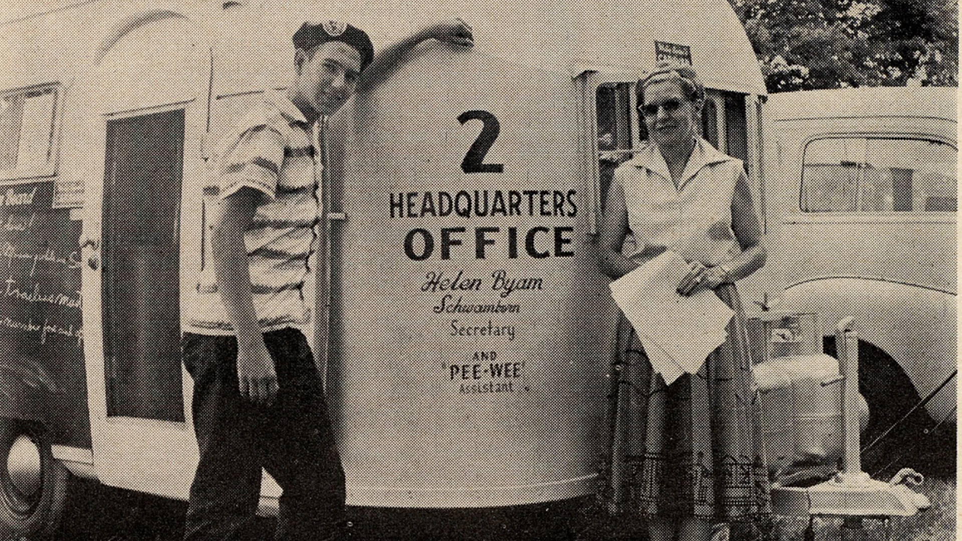 A young man and woman stand beside a trailer labeled "2 Headquarters Office," holding papers, with a vintage car in the background.