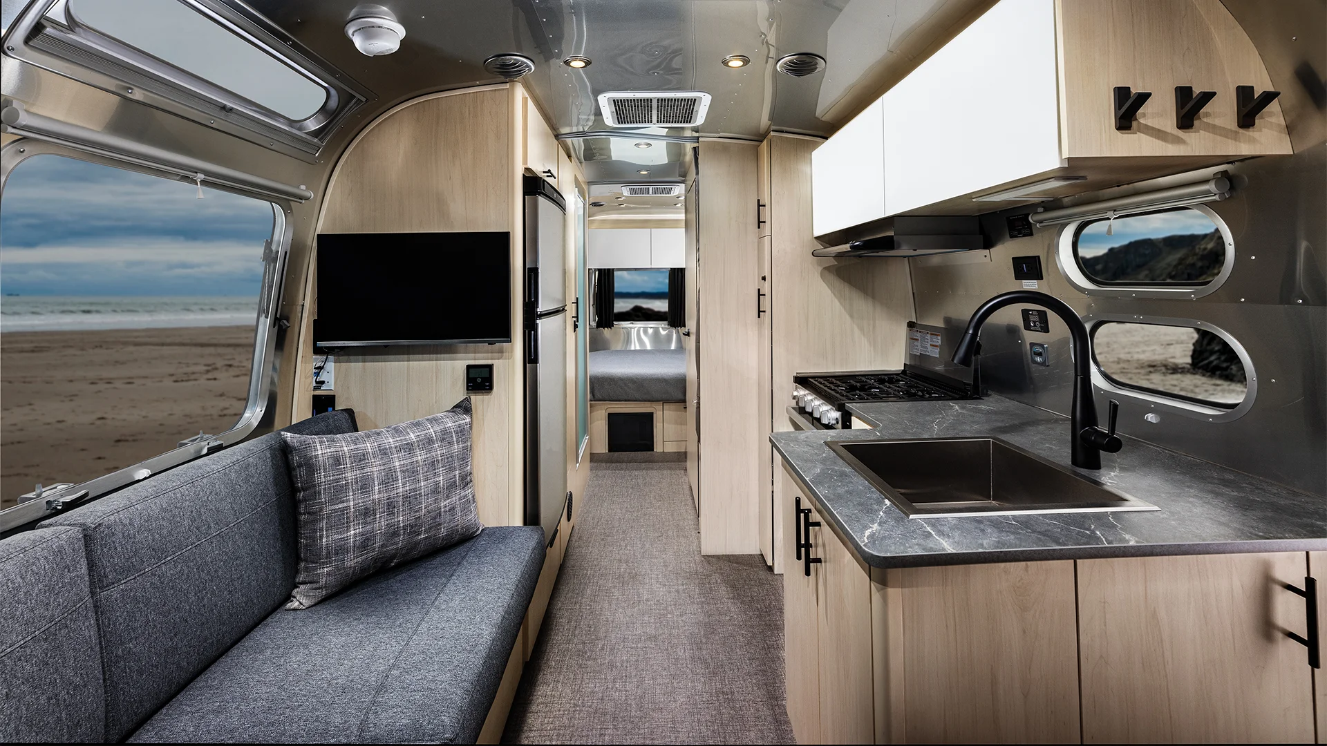 Interior of a modern RV with a kitchen, sofa, and TV. Large windows reveal a beach view. Light wood and stainless steel accents.