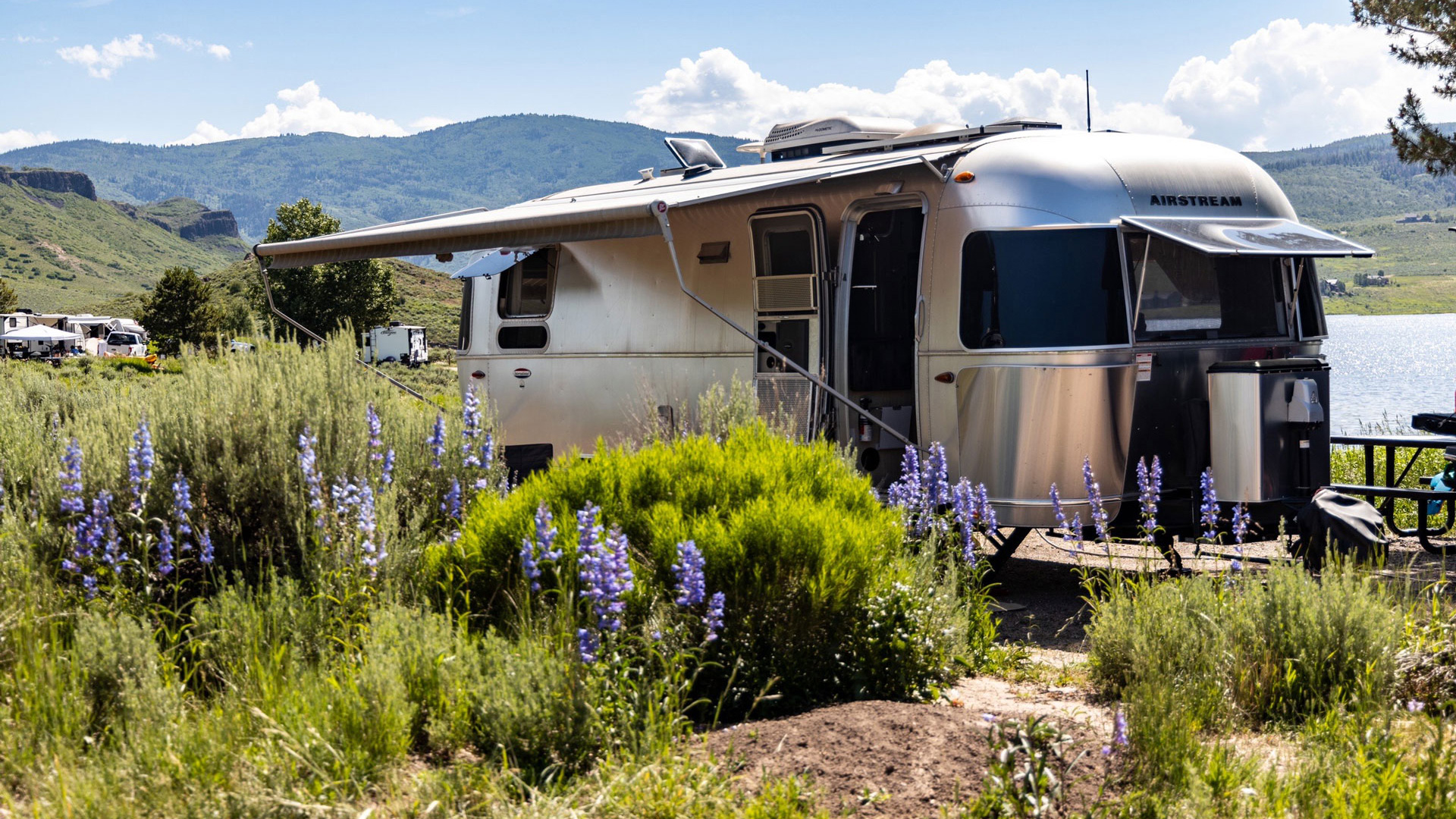 A silver Airstream trailer with an extended awning, parked near vibrant purple flowers, against a backdrop of mountains and a lake.