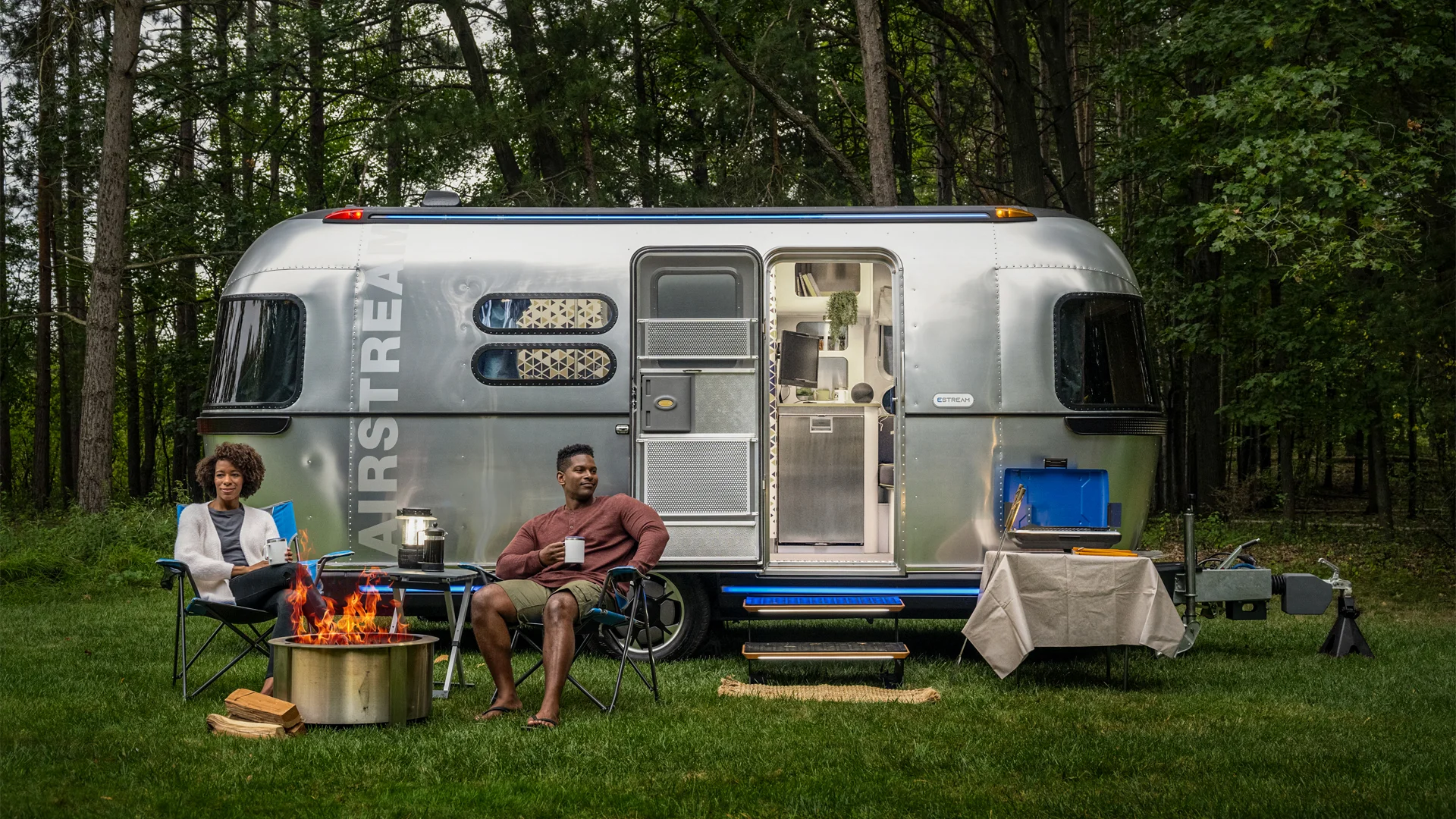 Two people sit by a campfire near a silver Airstream trailer in a forest setting, with camping equipment and a table nearby.