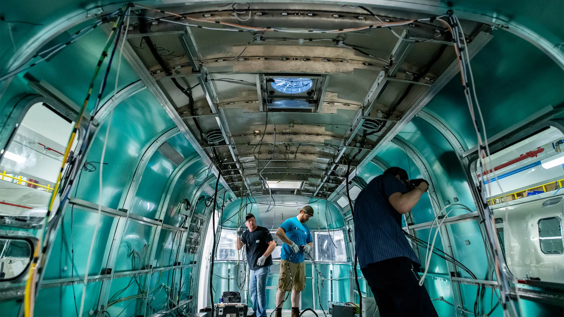 Three people inspect the interior of an aircraft under construction, with exposed wiring and green metallic walls.