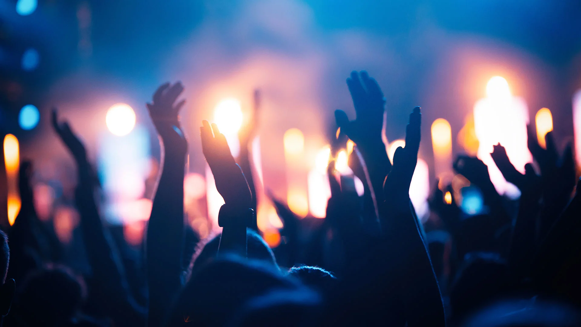 Silhouetted crowd with raised hands at a vibrant concert, illuminated by colorful stage lights.