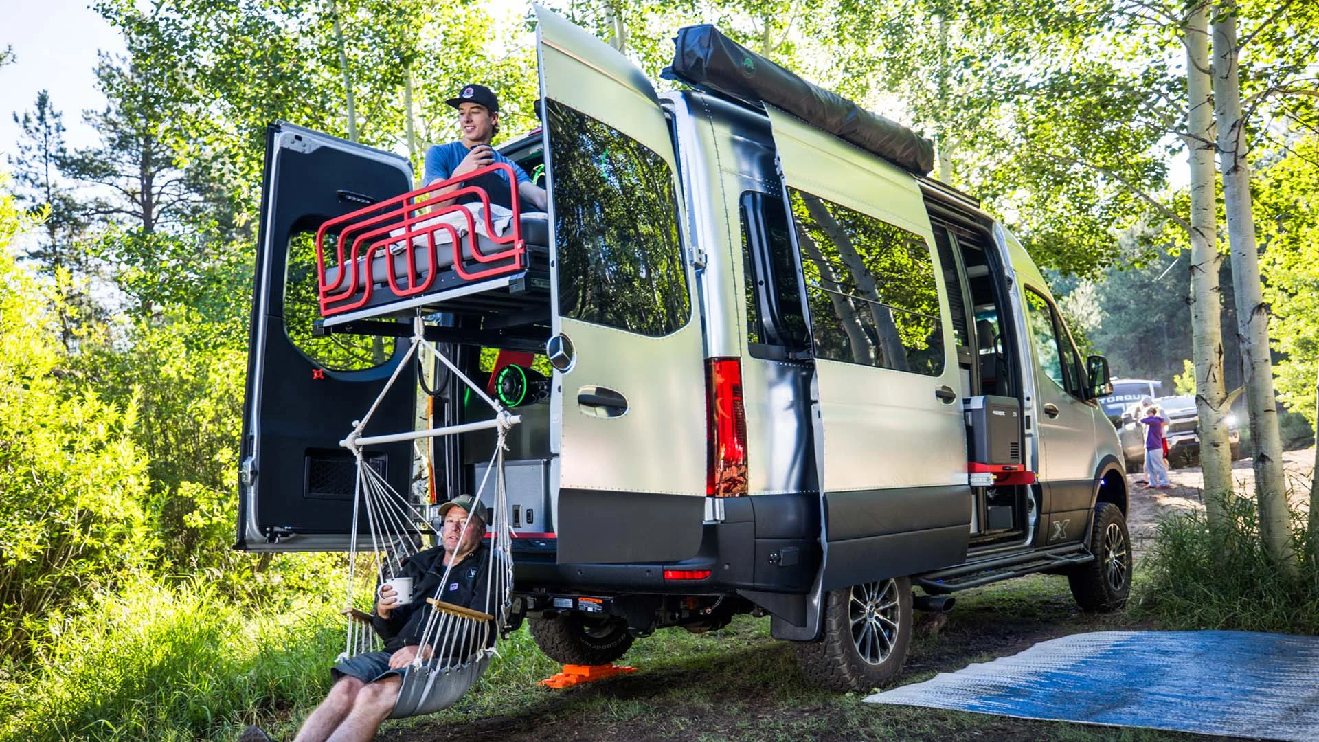 A camper van parked in a forest, with one person sitting on the open rear door and another relaxing in a hammock attached to the van.