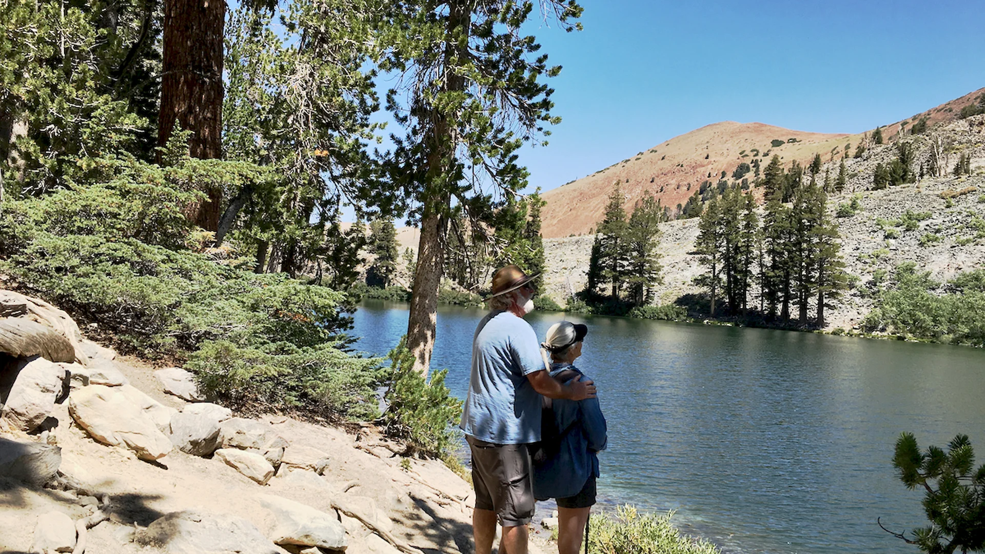 A couple wearing hats enjoys a serene view of a lake surrounded by trees and mountains on a sunny day.