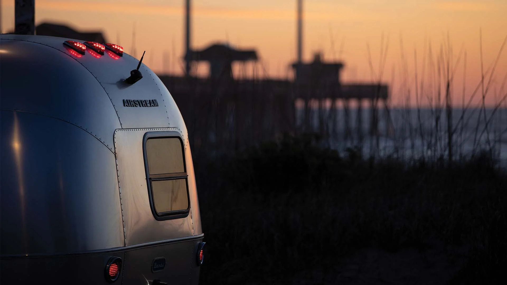 Airstream trailer parked by the beach at sunset, with a blurred pier and ocean in the background.