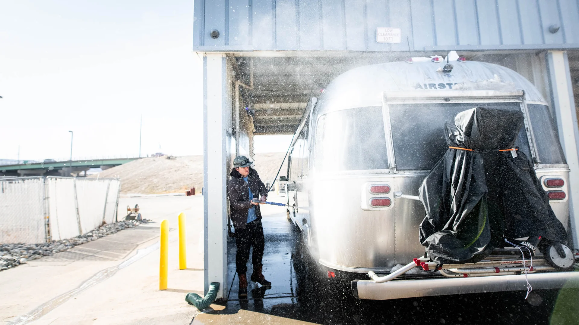 A person uses a hose to wash a silver Airstream trailer at an outdoor washing station, with water spraying and sunlight creating a bright scene.