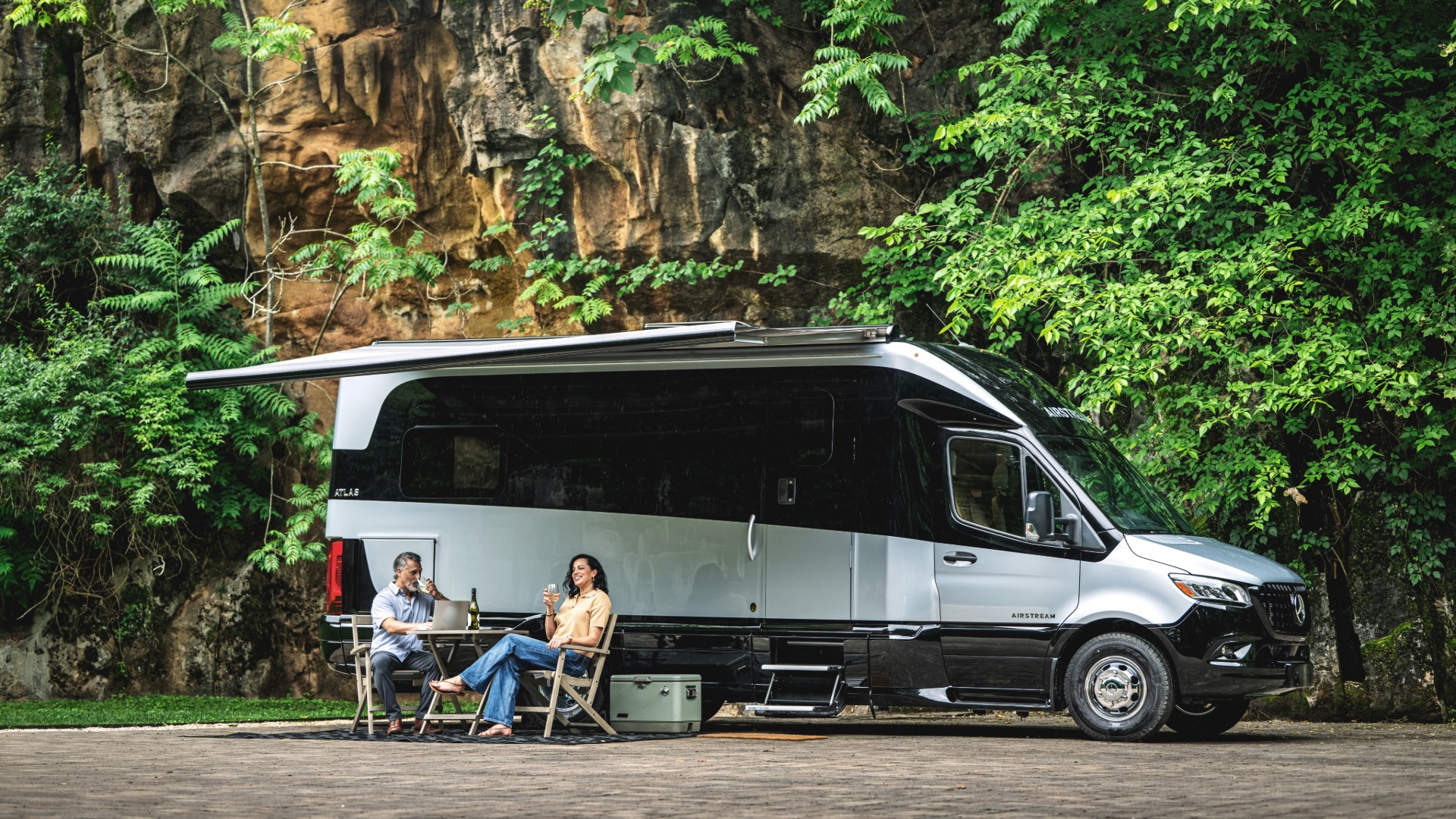 A couple sits under an awning, enjoying drinks beside a parked camper van amidst lush greenery and a rocky backdrop.