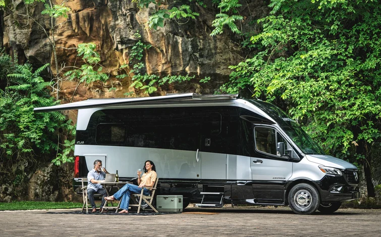 A couple sits under an awning, enjoying drinks beside a parked camper van amidst lush greenery and a rocky backdrop.