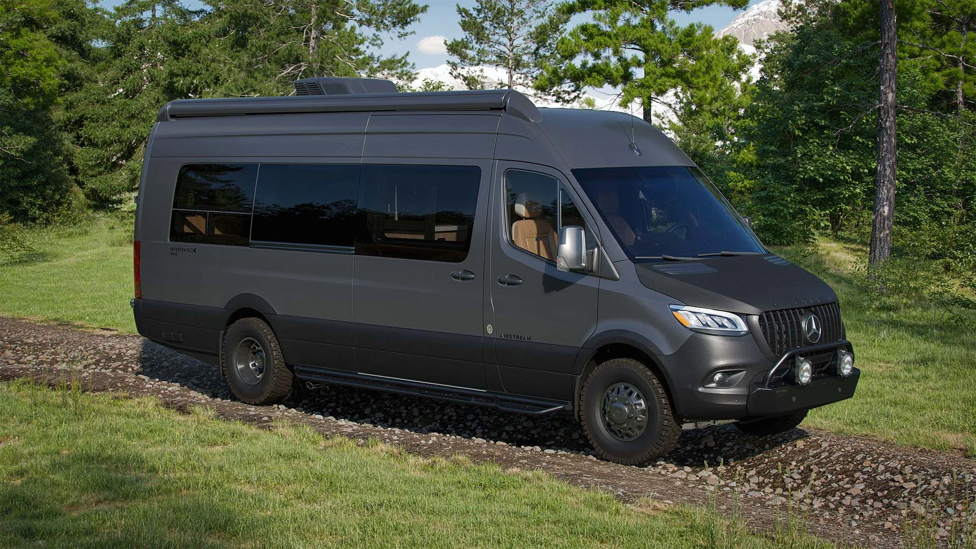 Gray camper van on a rocky path, surrounded by lush green trees and grass, under a clear sky.