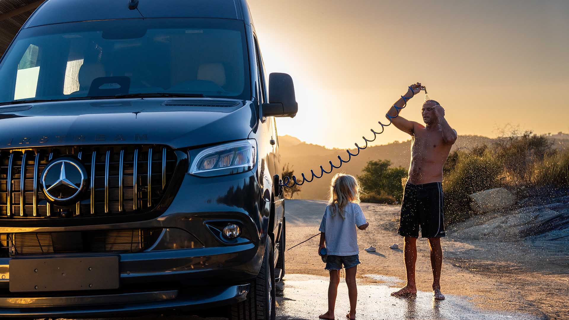 A man showers outside next to a black van in the sunset, while a child watches. Desert landscape in the background.