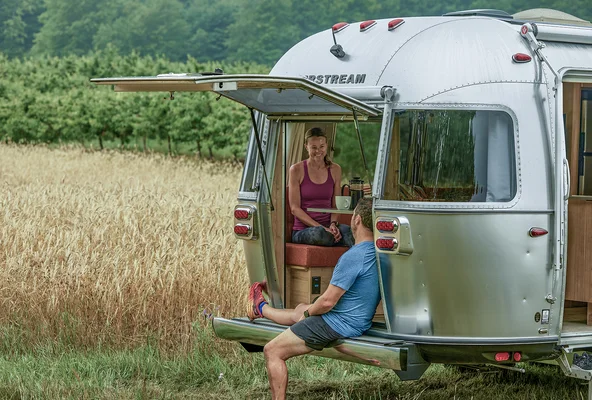 A woman sits inside the open rear of a silver Airstream camper, smiling at a man sitting on the back edge. They are parked in a grassy field near trees under an overcast sky.