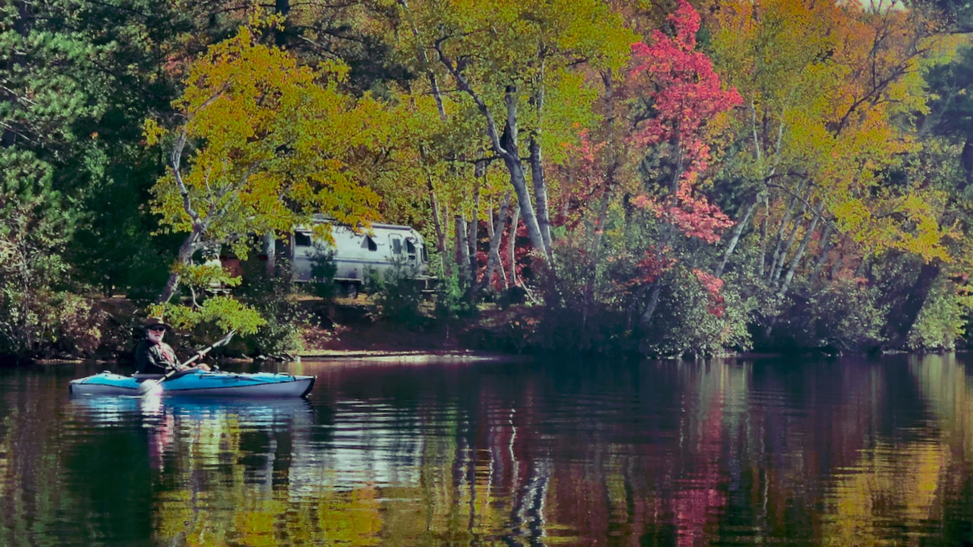 A person in a blue kayak paddles on a calm lake, surrounded by trees with vibrant autumn foliage. A white camper is parked among the trees on the shore in the background.