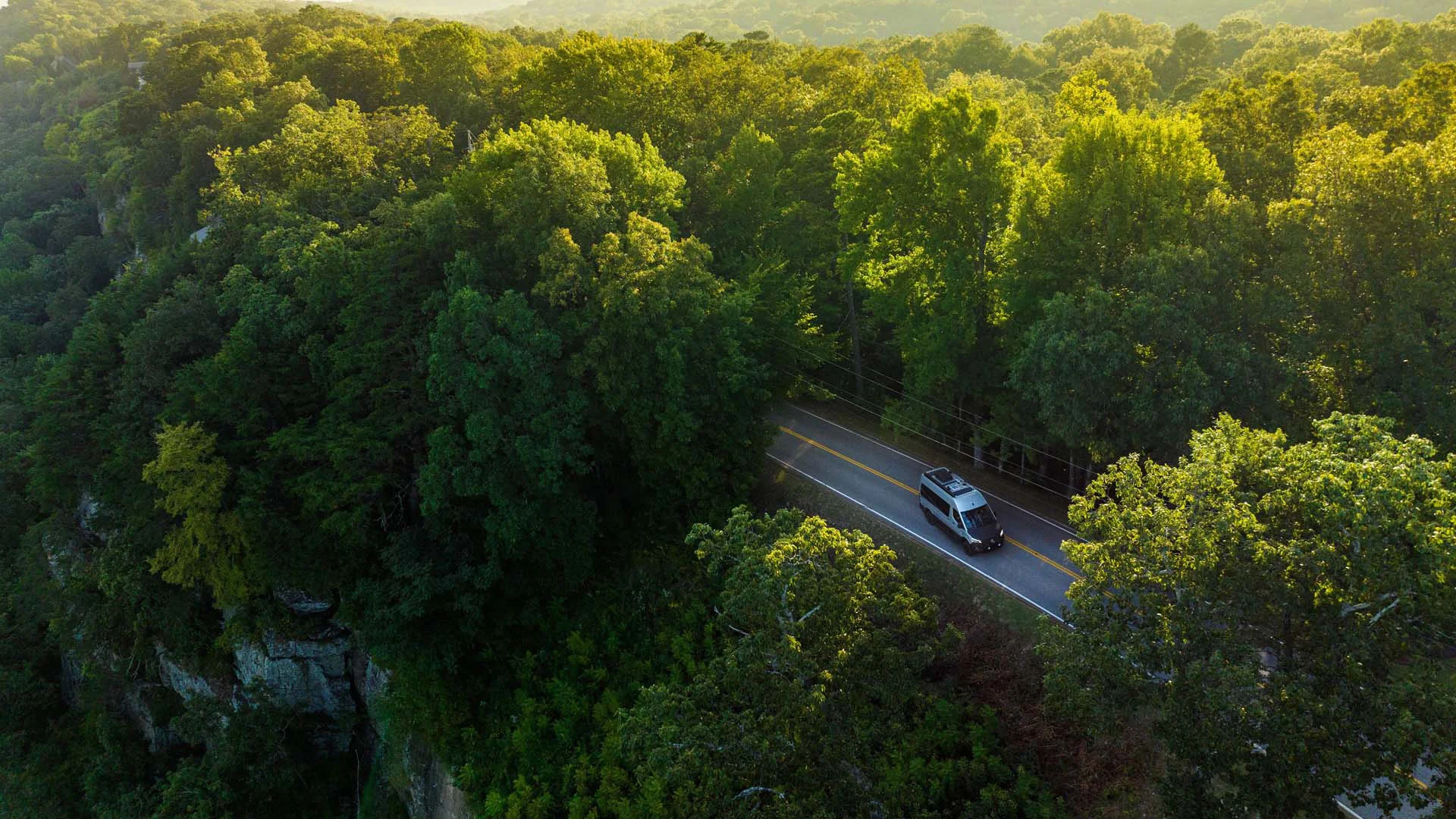 Aerial view of a car driving on a winding road through a lush, dense forest under bright sunlight.