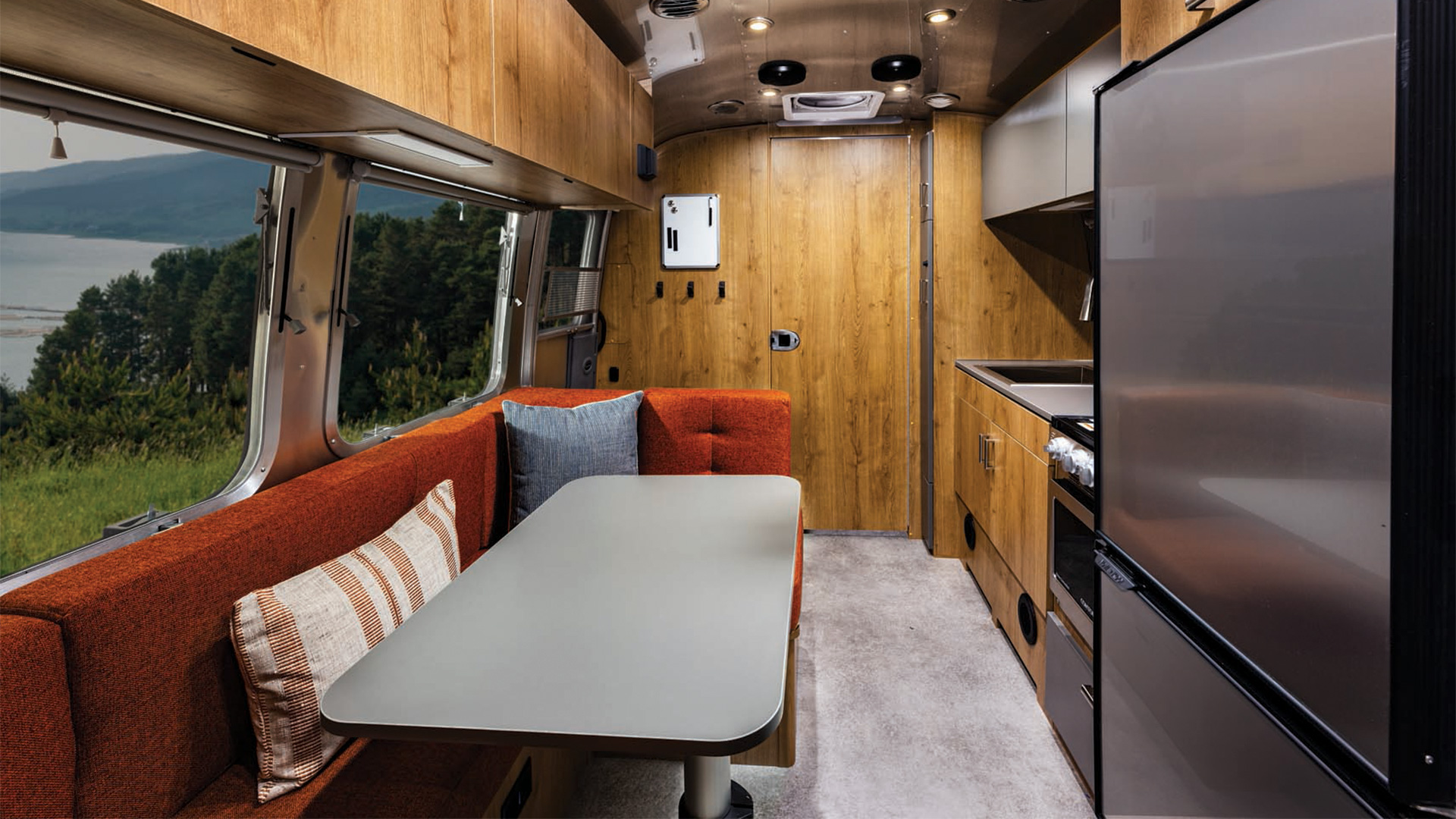 Interior of a modern camper van with wooden panels, a red cushioned bench with pillows, a sleek kitchen area, and a scenic window view.