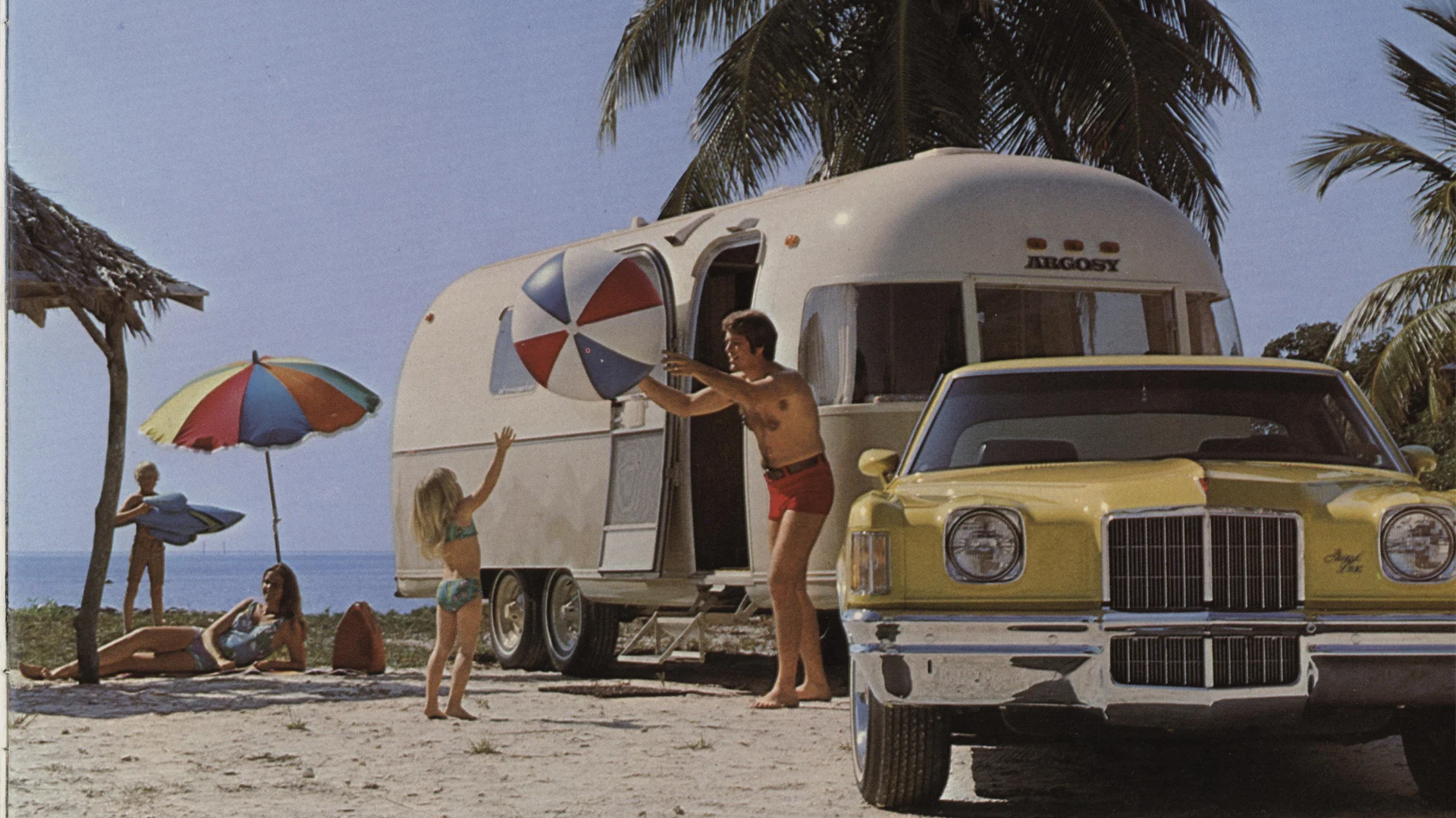 A man in swim trunks hands a beach ball to a child near a silver travel trailer and a yellow vintage car on a sunny beach, with palm trees, umbrellas, and people relaxing in the background.