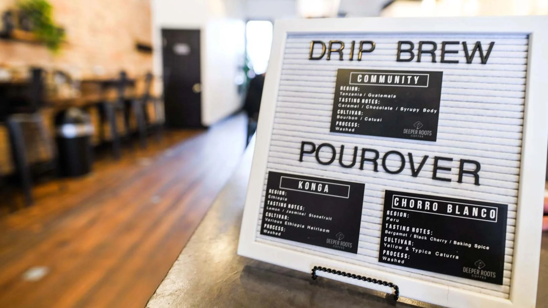 Menu board in a cafe displaying drip brew and pourover coffee options, with blurred seating area and counter in the background.
