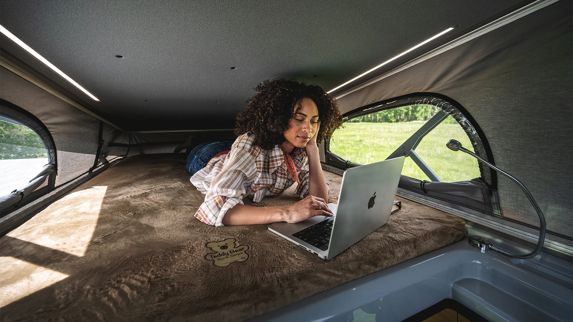 Woman working on a laptop inside a cozy camper van with a cushioned interior, natural light entering through side windows.