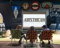 Three people sit on woven chairs watching a screen in the Airstream Heritage Center, surrounded by retro camping memorabilia and posters.