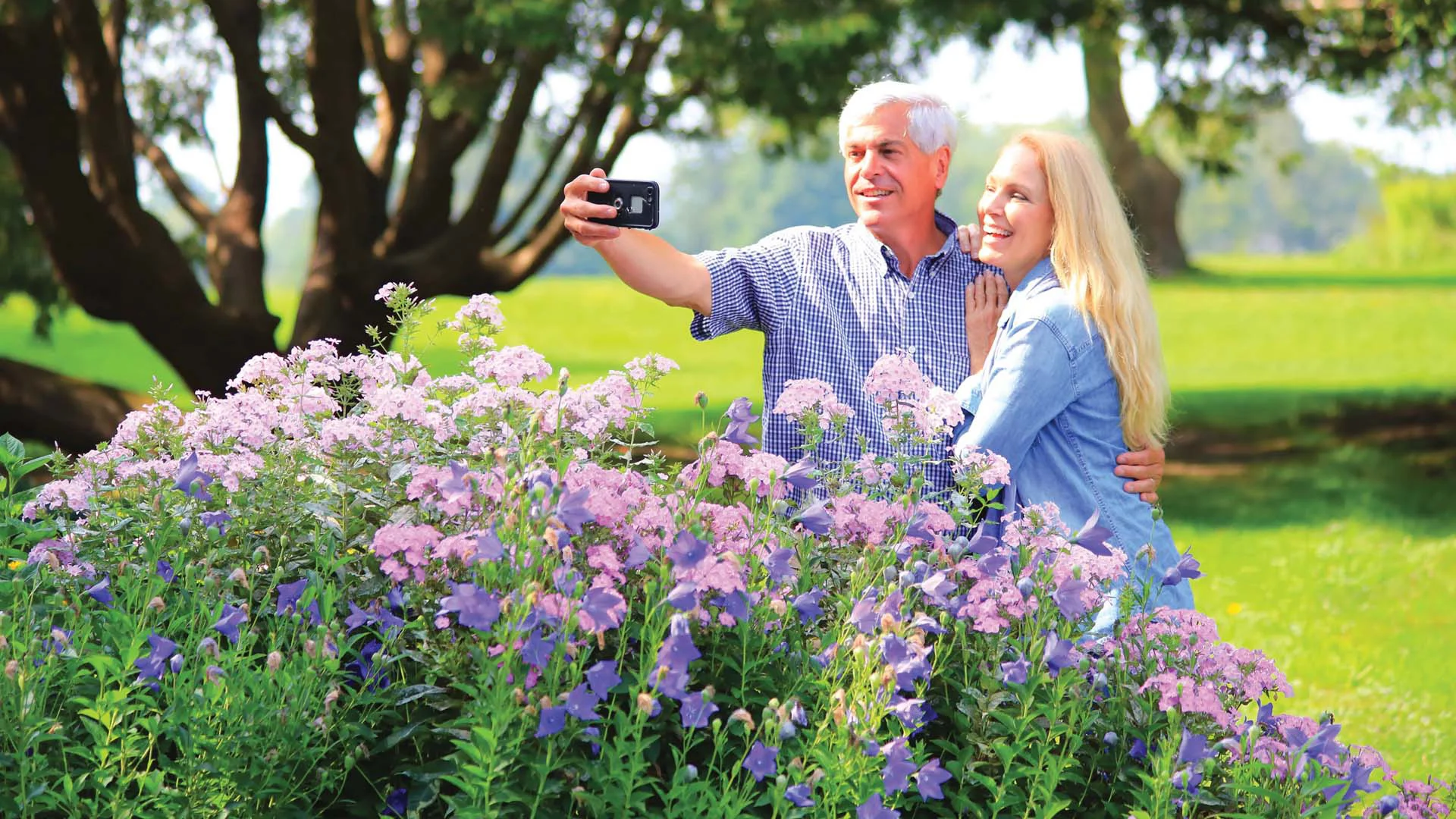 A smiling couple takes a selfie in a garden, surrounded by lush, blooming purple and pink flowers on a sunny day.