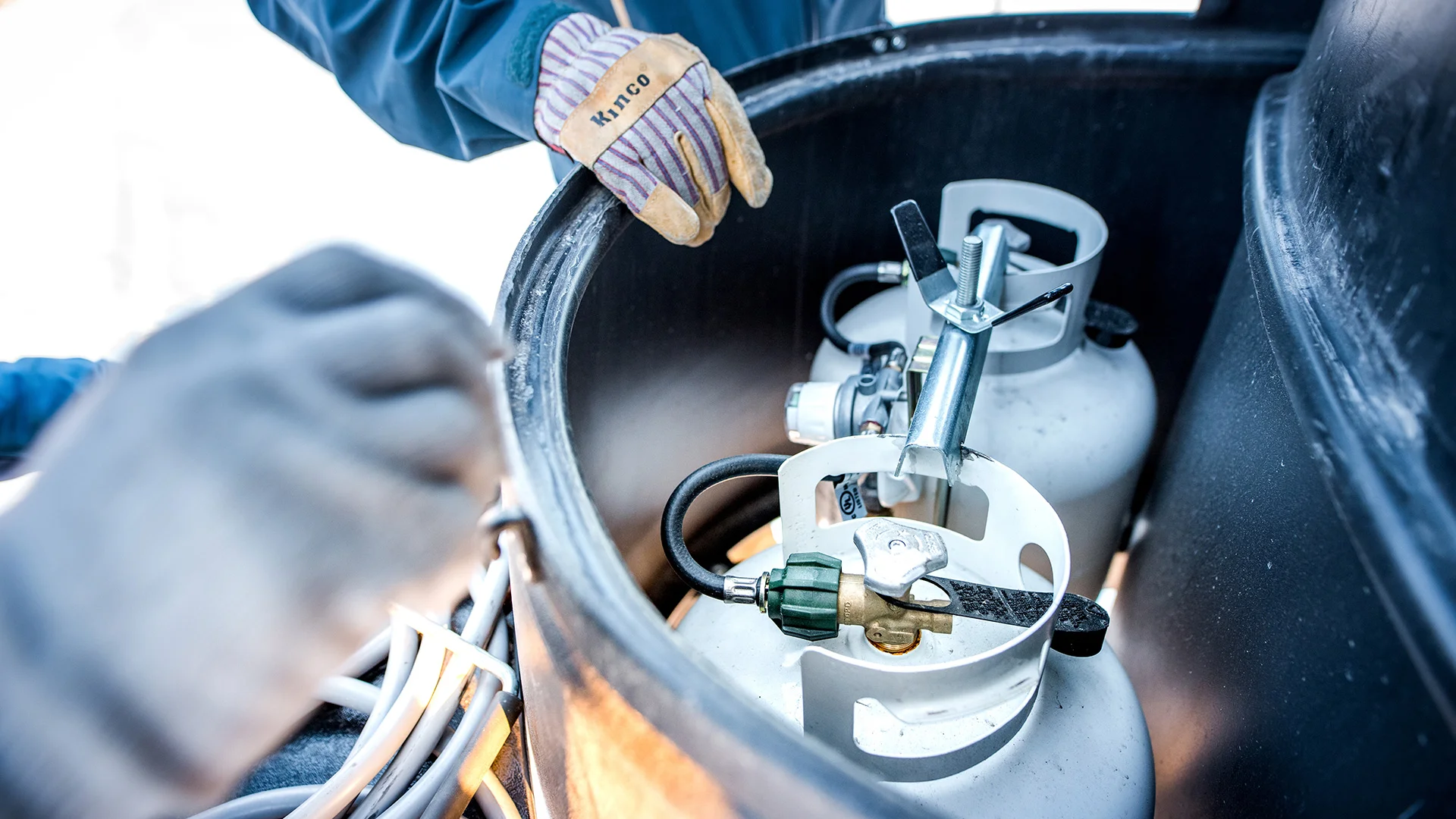 Two people handle propane tanks inside a black container. One person wears gloves labeled "HSI100." Hoses and connectors are visible.