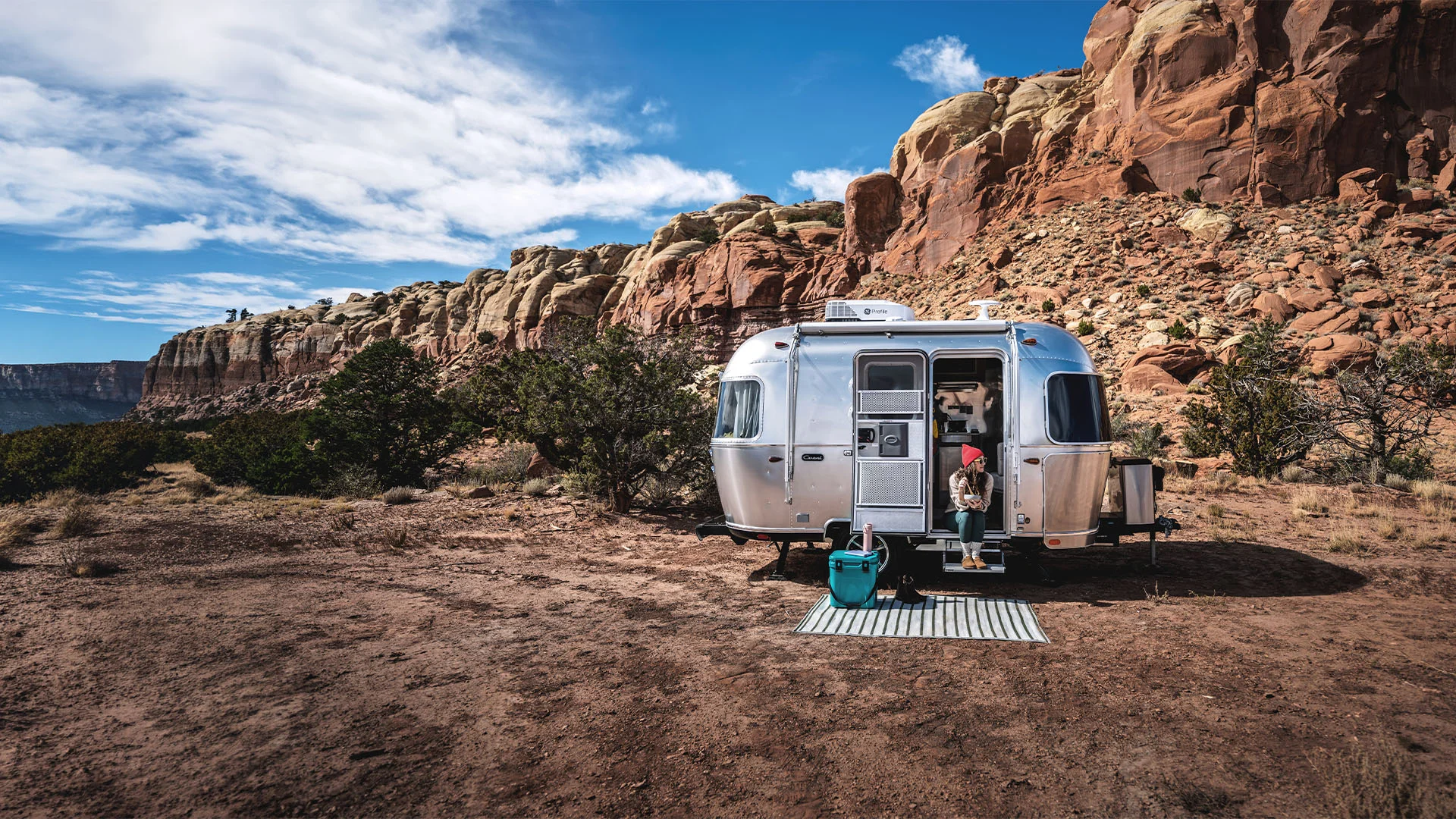 A silver Airstream trailer is parked on dry, rocky land near red cliffs and sparse bushes under a blue sky with clouds. A person sits at the entrance, enjoying the scenic desert landscape.