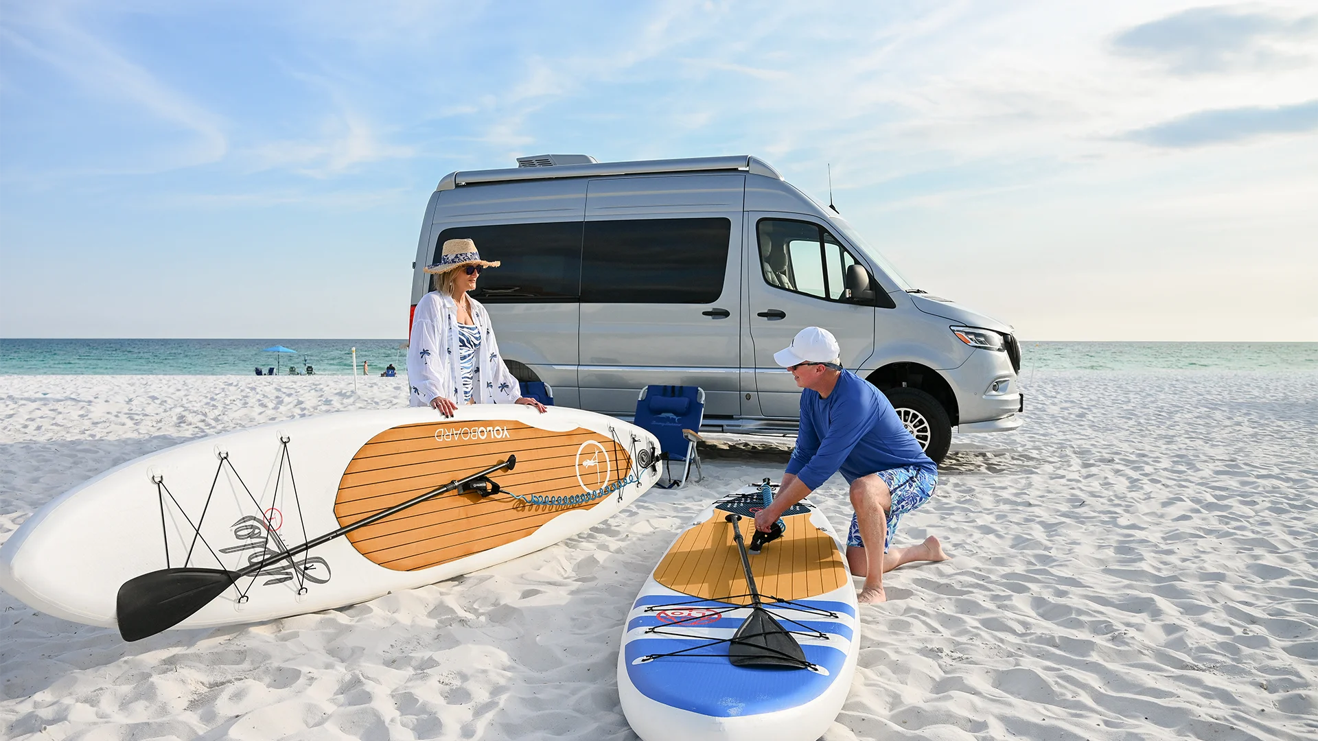 A man and woman with paddleboards stand near a camper van on a sandy beach, with the ocean in the background.