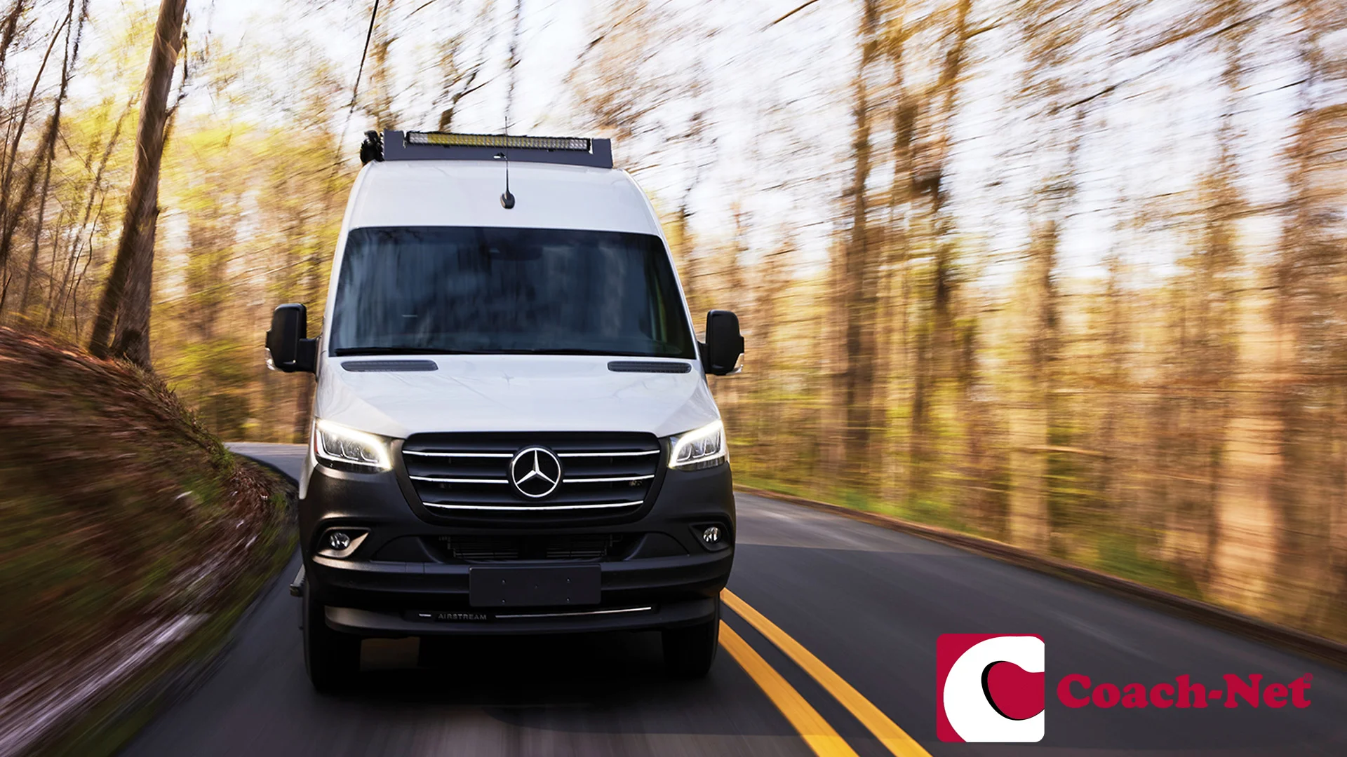 White Mercedes-Benz van driving on a winding road in a forest, with blurred trees in the background. Coach-Net logo in the corner.