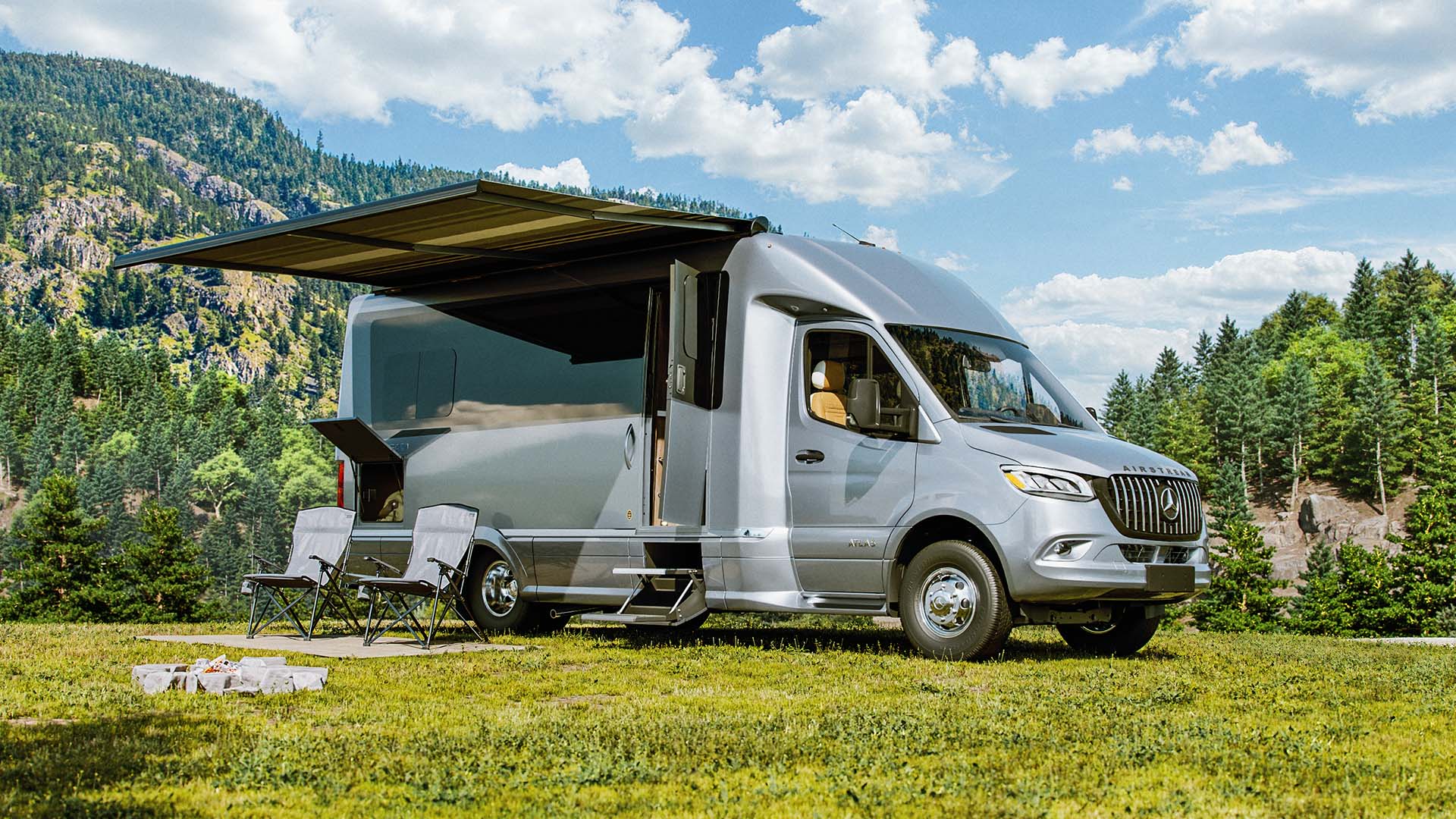 Silver camper van with an extended awning, parked on grass, surrounded by chairs and scenic mountains under a blue sky.