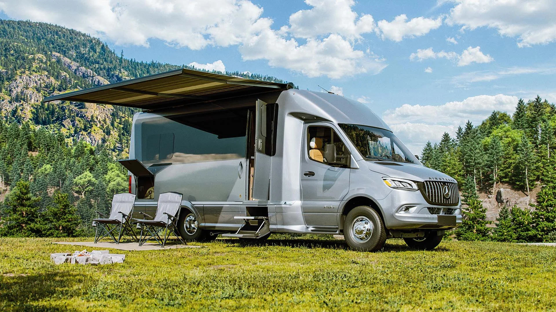 Silver camper van with an extended awning, parked on grass, surrounded by chairs and scenic mountains under a blue sky.