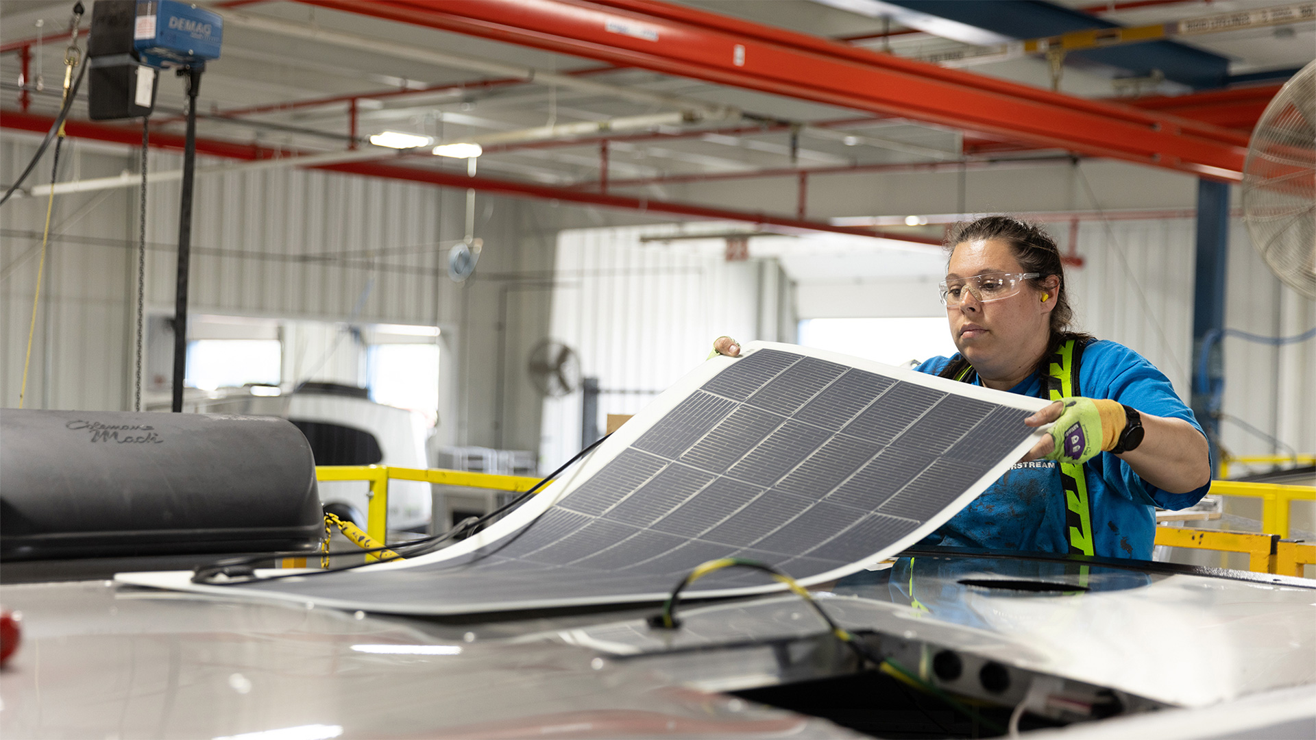 Person wearing safety gear installing a solar panel on a vehicle roof inside an industrial facility.