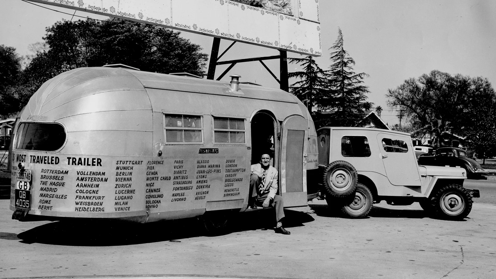 A vintage black-and-white photo shows a man sitting in the doorway of a shiny metal travel trailer hitched to a Jeep. The trailer displays names of European cities visited, and trees and a billboard are in the background.