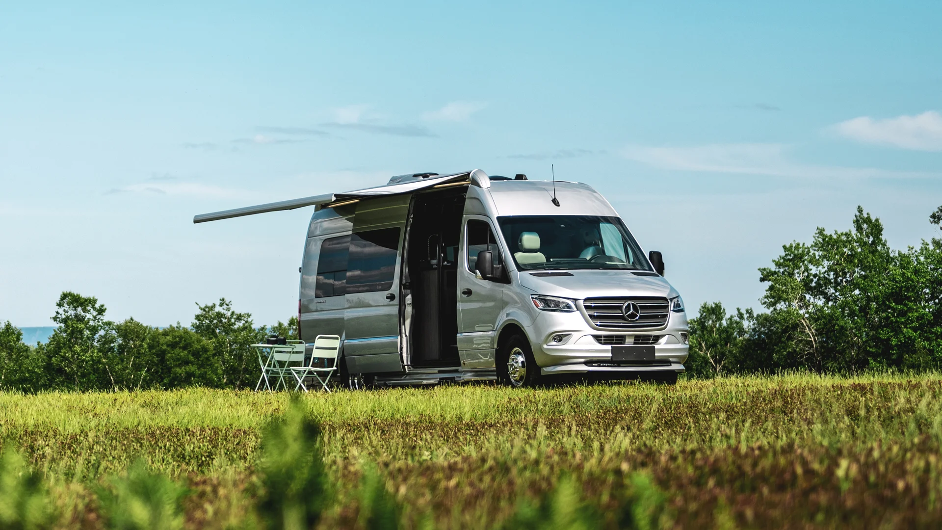 Silver camper van with an extended awning parked on a grassy field, surrounded by trees. A small table and chairs are set up nearby.