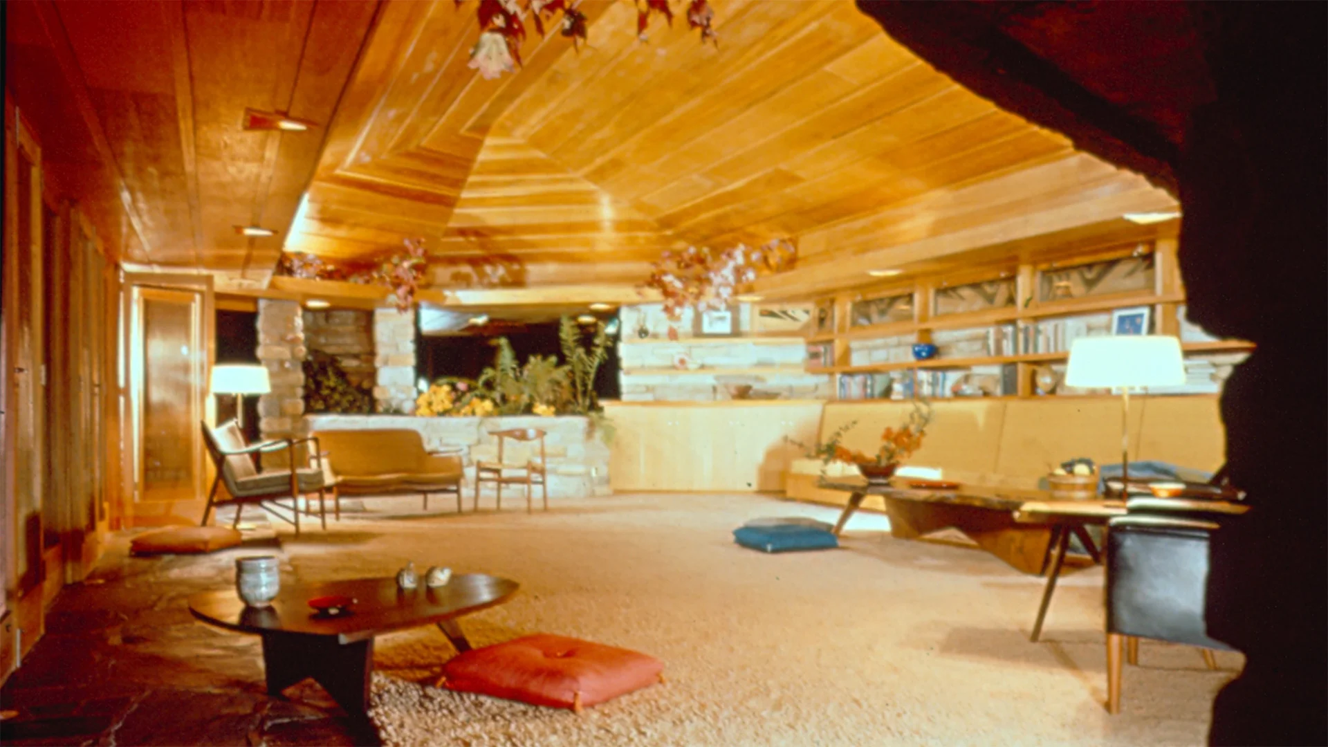 Spacious mid-century modern living room with wooden ceiling, plush carpet, cushions, low tables, chairs, and a wall of shelves with decor.