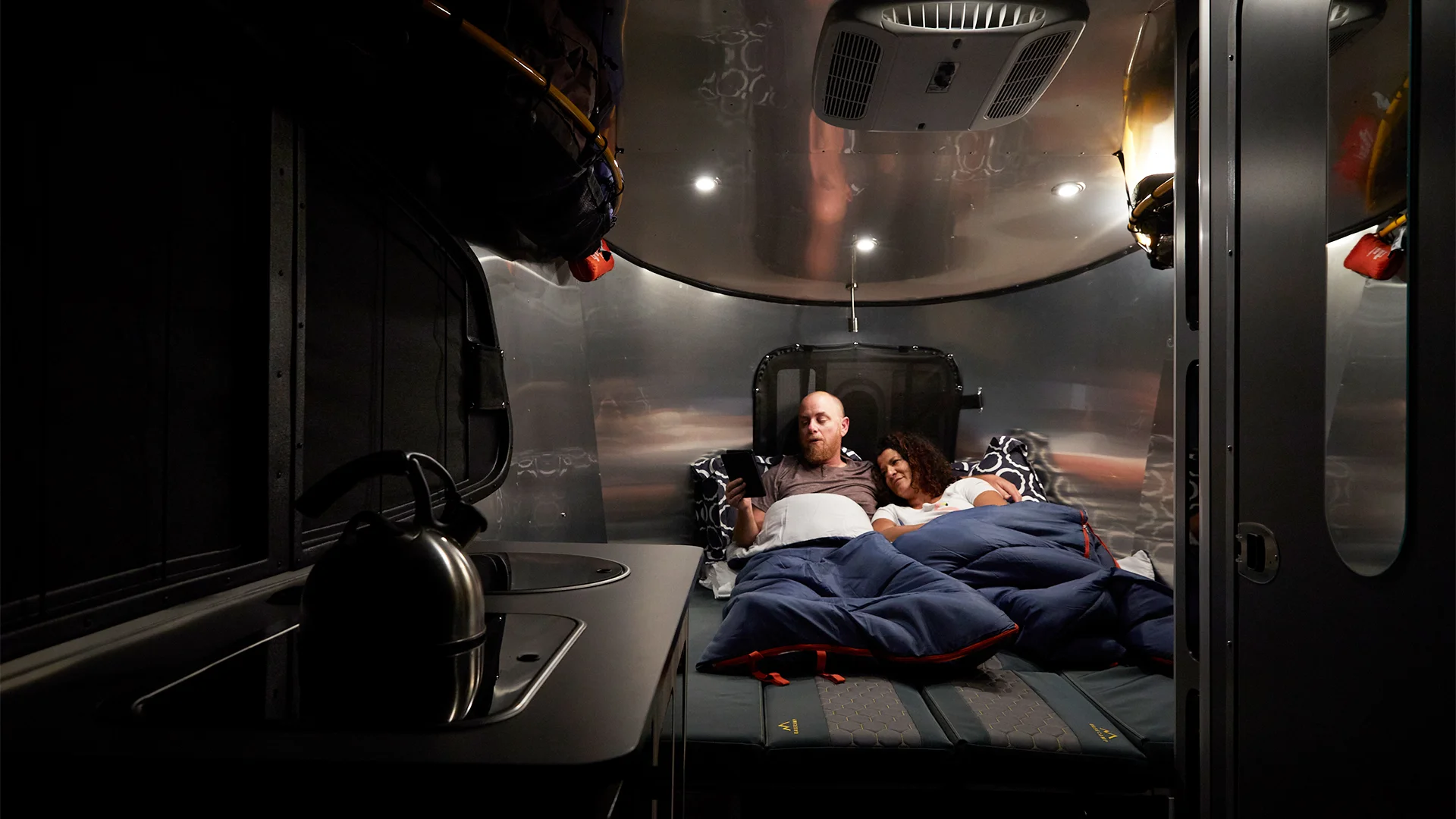 A couple sleeps in sleeping bags on a bed inside a modern camper van, with a compact kitchenette featuring a kettle nearby.