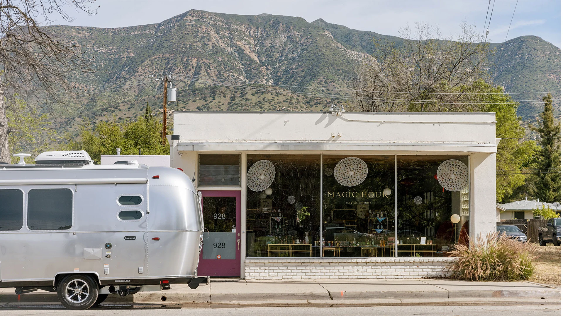 Storefront with large windows displaying decorative items, white building, parked silver trailer, mountains in the background.