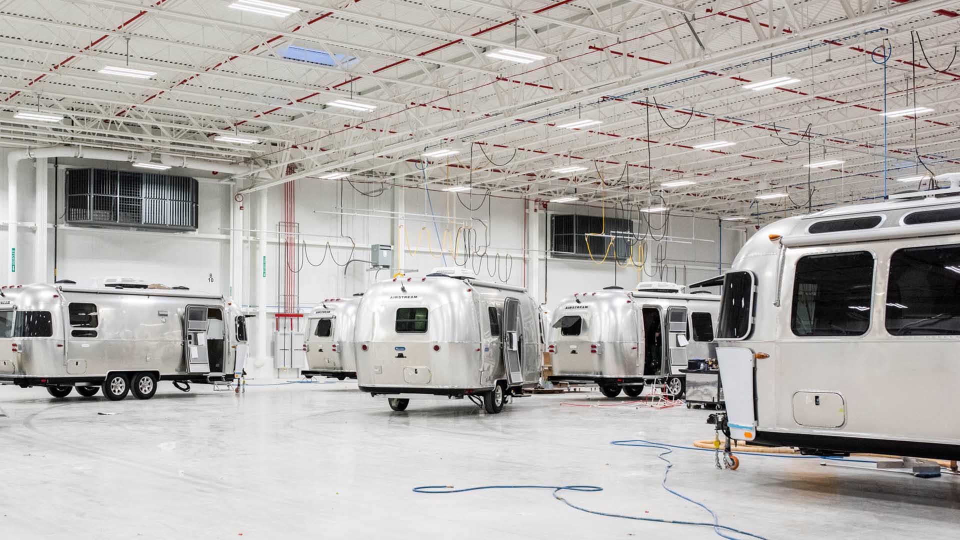 Several silver Airstream trailers inside a spacious factory with high ceilings and overhead cables.