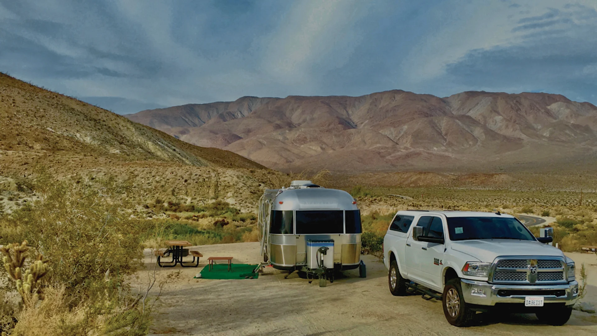 A silver Airstream trailer and a white pickup truck are parked at a desert campsite, surrounded by dry bushes, with mountains rising in the background under a partly cloudy sky.